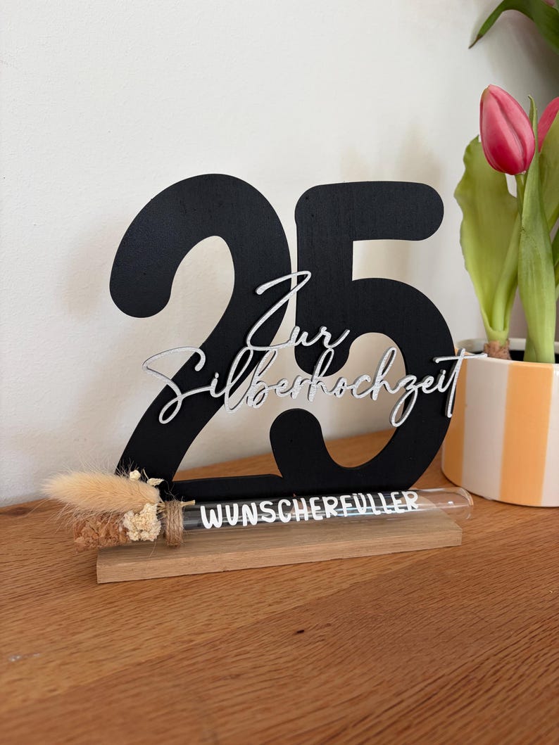 May include: A black wooden '25' centerpiece with the words 'Zur Silberhochzeit' in silver script. A small glass tube with the word 'WUNSCHERFUELLER' sits on a wooden base, with dried flowers.