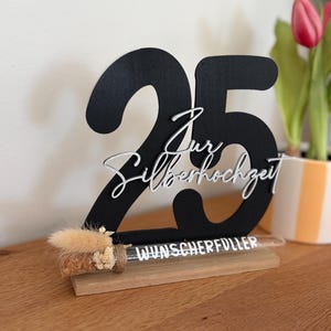 May include: A black wooden table decoration with the number 25 and the words "Zur Silberhochzeit" in white script. A small glass tube with dried flowers and the word "Wunscherfuller" is attached to the base. The decoration is on a wooden surface.