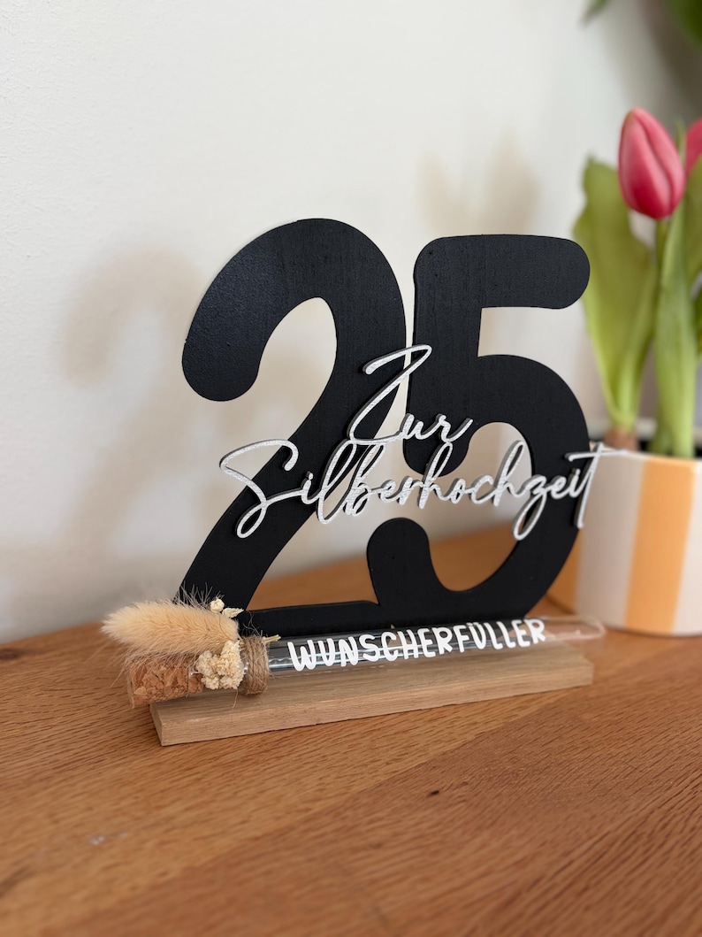 May include: A black wooden table decoration with the number 25 and the words "Zur Silberhochzeit" in white script. A small glass tube with dried flowers and the word "Wunscherfuller" is attached to the base. The decoration is on a wooden surface.