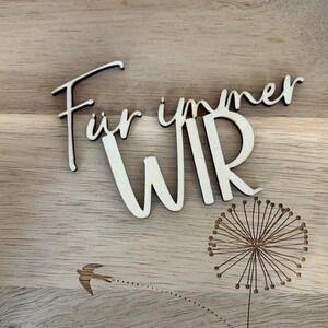 May include: Wooden plaque with the German words "F&uuml;r immer WIR" (Forever US) in a cursive font. A dandelion and a flying bird are etched into the wood. The plaque is made of light-colored wood.