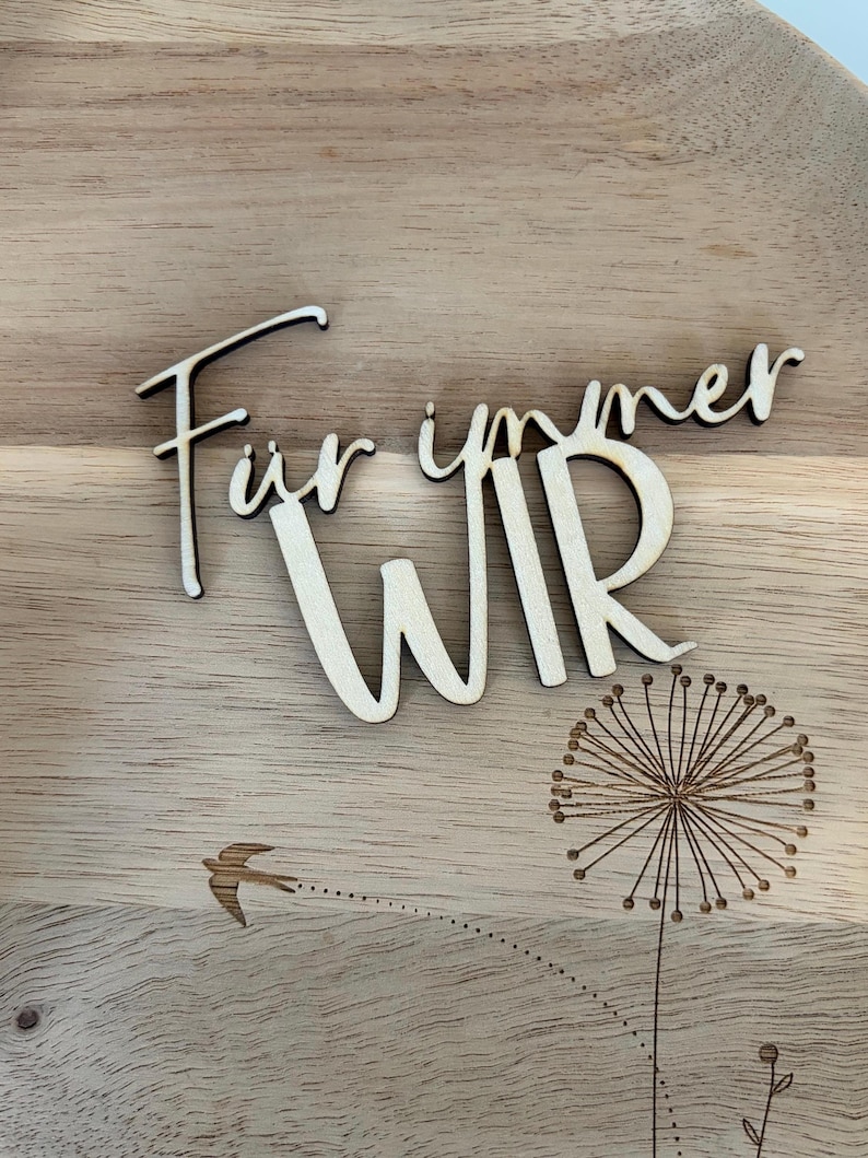 May include: Wooden plaque with the German words "F&uuml;r immer WIR" (Forever US) in a cursive font. A dandelion and a flying bird are etched into the wood. The plaque is made of light-colored wood.