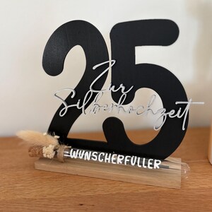 May include: A black wooden number 25 with the words "Zur Silberhochzeit" in silver script. The base includes a wooden stand with the word "WUNSCHERF&Uuml;LLER" and a small dried floral arrangement. The item is on a wooden surface.
