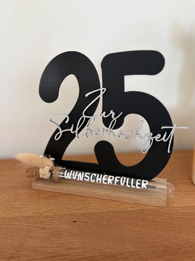 May include: A black wooden number 25 with the words "Zur Silberhochzeit" in silver script. The base includes a wooden stand with the word "WUNSCHERF&Uuml;LLER" and a small dried floral arrangement. The item is on a wooden surface.