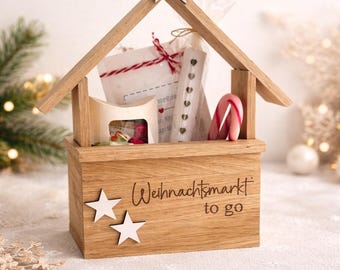 May include: A light brown wooden house-shaped box with a peaked roof and a star on top. The front is engraved with the text "Weihnachtsmarkt to go" and decorated with two white stars. Inside are small gift bags, a candy cane, and a box.