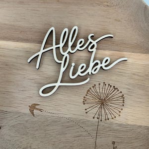 May include: Wooden decorative tray with the German phrase "Alles Liebe" in cursive script. The tray features a laser-etched dandelion and a small bird in flight, all set against a light wood grain background.