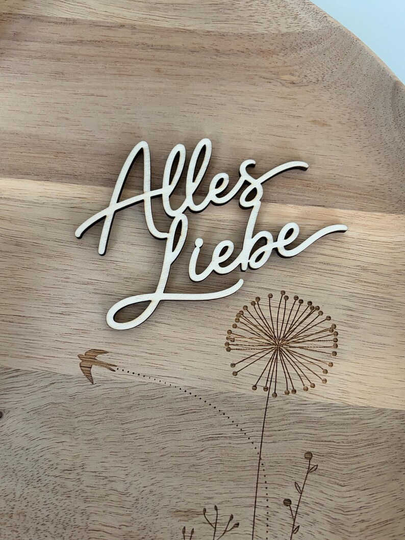 May include: Wooden decorative tray with the German phrase "Alles Liebe" in cursive script. The tray features a laser-etched dandelion and a small bird in flight, all set against a light wood grain background.