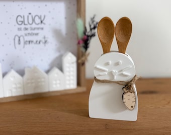 May include: A white ceramic bunny-shaped utensil holder with two wooden spoons inserted as ears. A wooden carrot charm hangs from a twine bow around the bunny's neck. A framed picture with the text "Glück ist der Summe schöner Momente" is in the background.
