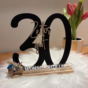 May include: A black wooden '30' birthday decoration with the words "Zeitos gut" in a lighter color. A small glass tube with text is attached to a wooden base, decorated with dried flowers.