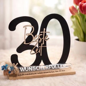 May include: A black wooden '30' centerpiece with the words 'Beste Zeit' in a light wood script. A glass tube with the word 'WUNSCHERF&Uuml;LLER' is on a wooden base, with a small floral arrangement.