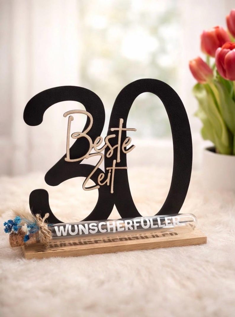 May include: A black wooden '30' centerpiece with the words 'Beste Zeit' in a light wood script. A glass tube with the word 'WUNSCHERF&Uuml;LLER' is on a wooden base, with a small floral arrangement.
