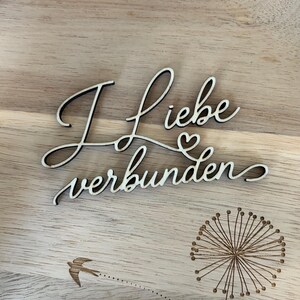 May include: Wooden sign with the German phrase "I love connected" in a cursive font. The sign also features a dandelion and a bird illustration, all etched into the wood. The background is a light brown wooden surface.