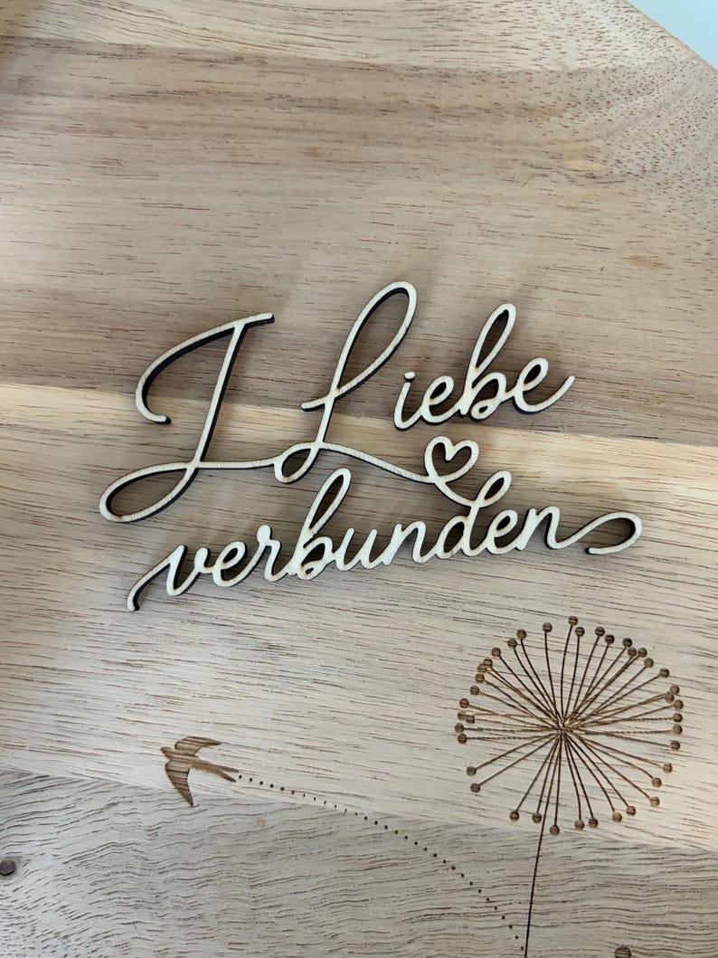 May include: Wooden sign with the German phrase "I love connected" in a cursive font. The sign also features a dandelion and a bird illustration, all etched into the wood. The background is a light brown wooden surface.