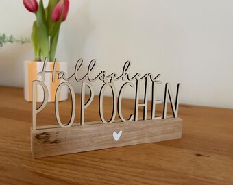 May include: Wooden sign with the word "Hallöchen POPOCHEN" in a playful font. The sign is made of light-colored wood with black outlines and sits on a wooden base with a white heart. A vase with pink tulips is in the background.