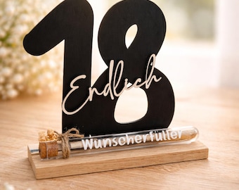 May include: A black wooden number 18 with the word "Endlich" in a cursive font, and a glass tube with the words "Wunscherfüller" on a wooden base. The tube is sealed with a cork and twine.