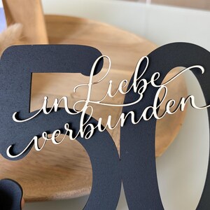 May include: A black wooden number 50 with the words "Liebe in verbunden" in a cursive font. The number is cut out, and the text is layered on top. The background includes a wooden tray and a white vase with greenery.
