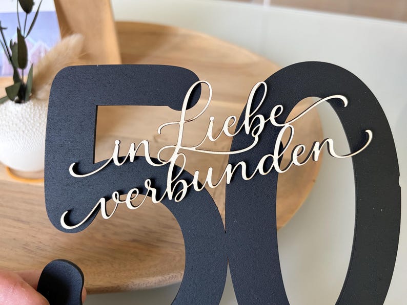 May include: A black wooden number 50 with the words "Liebe in verbunden" in a cursive font. The number is cut out, and the text is layered on top. The background includes a wooden tray and a white vase with greenery.