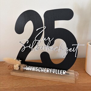May include: A black wooden decoration with the number 25, and the words "Zur Silberhochzeit" in silver script. A small glass tube with the words "Wunscherf&uuml;ller" sits on a wooden base, with dried flowers tied to it. The decoration is on a wooden surface.