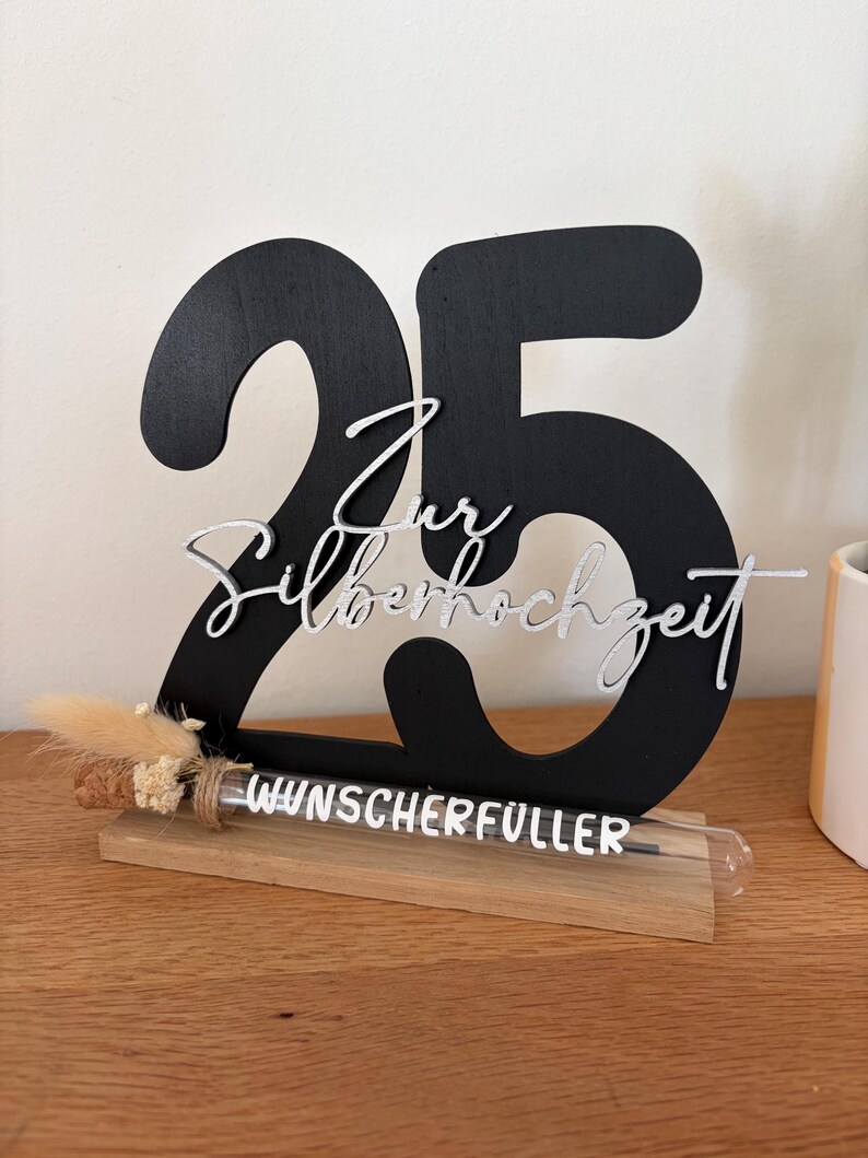 May include: A black wooden decoration with the number 25, and the words "Zur Silberhochzeit" in silver script. A small glass tube with the words "Wunscherf&uuml;ller" sits on a wooden base, with dried flowers tied to it. The decoration is on a wooden surface.