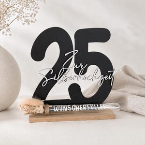 May include: A black wooden number 25 with the words "Zur Silberhochzeit" in white script. A small glass tube with the word "WUNSCHERF&Uuml;LLER" is attached to a wooden base. A vase with dried flowers is in the background.