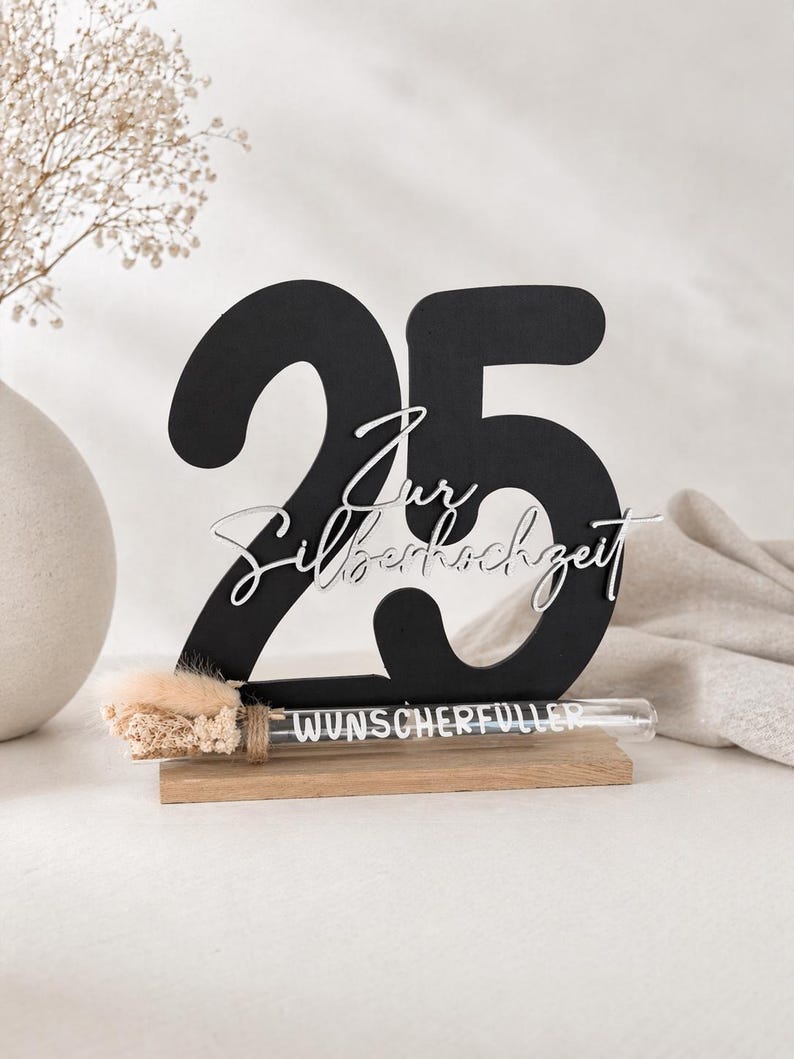 May include: A black wooden number 25 with the words "Zur Silberhochzeit" in white script. A small glass tube with the word "WUNSCHERF&Uuml;LLER" is attached to a wooden base. A vase with dried flowers is in the background.
