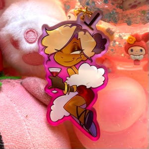 Puede incluir: Un colgante acrílico transparente con un personaje de dibujos animados con cabello rubio, un cuello de piel blanco y un vestido rojo, sosteniendo una copa de vino tinto. El colgante tiene un borde rosa y un llavero dorado.