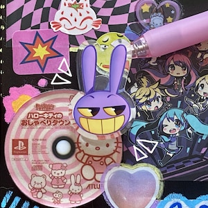 Può includere: Una collezione di adesivi colorati e un CD sono disposti su una superficie nera. Gli adesivi includono un coniglio viola con un sorriso giallo, una stella e un gatto fragola. C'è anche una penna rosa.