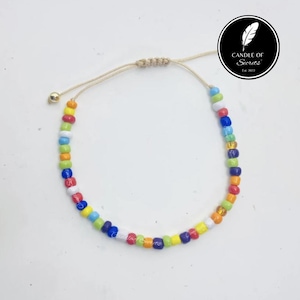 Peut inclure: Un bracelet de perles multicolores avec une fermeture à nœud coulissant. Le bracelet est composé de petites perles rondes aux couleurs de l'arc-en-ciel. Le bracelet est sur une surface blanche. Le logo "Candle of Secrets" est en haut à droite.