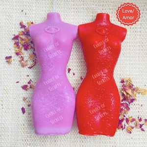 May include: Two hourglass-shaped candles in the form of a female torso. The candle on the left is pink and the candle on the right is red. Both candles have the text "Candle of Secrets" printed on them.