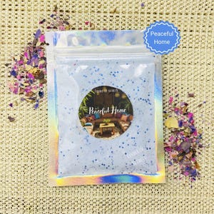 May include: A holographic pouch containing blue glitter and a circular label with the words "Peaceful Home." The label shows a living room scene. Dried flower petals in pink, purple, and yellow are scattered around the pouch.
