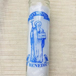 Puede incluir: Una vela de vidrio blanco con una imagen azul de San Benito. La vela tiene el texto "Candle of Secrets" y "San Benito" en ella.