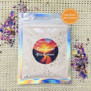 May include: A white pouch with a holographic iridescent finish contains a pink candle with green glitter. The candle has a round sticker with a sunset scene and the text "Open Roads/Abre Caminos" in black and white. The pouch has a smaller sticker with the text "Open Roads/Abre Caminos" in black and white.