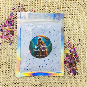May include: A small, clear plastic bag filled with a light blue powder with silver glitter. The bag has a holographic iridescent finish. A round sticker with a colourful image of a skeleton holding a scythe and the text "Holy Death! Santa Muerte Candle of Secrets" is on the front of the bag.