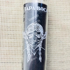 May include: A black glass candle with a white label that says "Candle of Secrets" and "Tapa Boca" with a drawing of a woman with her mouth covered. The bottom of the candle says "Shut Up" and "Candle of Secrets".