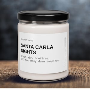 May include: A white candle in a glass jar with a black lid. The label reads "SANTA CARLA NIGHTS" with the text "ocean air, bonfires, and too many damn vampires." The candle is on a wooden surface.