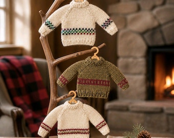 May include: Three miniature knitted sweaters hanging on a wooden branch display stand. The top sweater is cream with a multi-colored geometric pattern. The middle sweater is olive green with a maroon band. The bottom sweater is cream with a maroon band.
