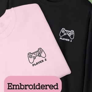 May include: Two embroidered sweatshirts, one pink and one black, with a video game controller design and the text "Player 1" and "Player 2".
