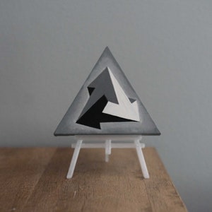 Può includere: Dipinto in scala di grigi di un triangolo con frecce, esposto su un piccolo cavalletto bianco. L'opera presenta un design geometrico con toni neri, bianchi e grigi, creando un effetto 3D. Lo sfondo è grigio neutro.