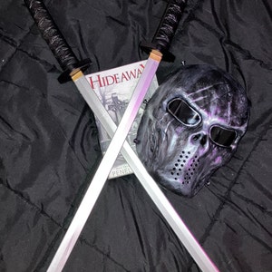 May include: Two silver-coloured swords with black handles are crossed over a book titled "Hideaway" with a black and grey skull mask on top of the book.