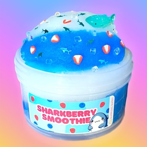 May include: A clear plastic container filled with blue and white slime, decorated with small shark and strawberry charms. The label reads "Sharkberry Smoothie" with a cartoon shark illustration. The slime is a vibrant, colorful product.