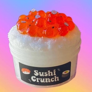 May include: A container of white slime topped with orange, translucent spheres and small flower-shaped decorations. The container has a black label with the words "Sushi Crunch" and sushi illustrations.