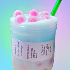 May include: A clear plastic cup filled with a light blue liquid, topped with white foam and pink boba pearls. A green straw is inserted into the drink. The label reads "Summer Skies" and "Dumpling Slime Shop".