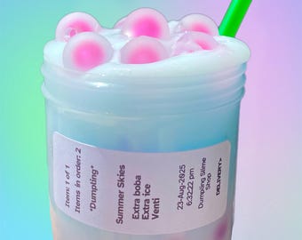 May include: A clear plastic cup filled with a light blue liquid, topped with white foam and pink boba pearls. A green straw is inserted into the drink. The label reads "Summer Skies" and "Dumpling Slime Shop".