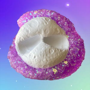 May include: A white, textured, moon-shaped object is surrounded by a sparkling, purple, glittery substance. The background transitions from purple to blue. Small yellow flower-shaped decorations are scattered around the object.