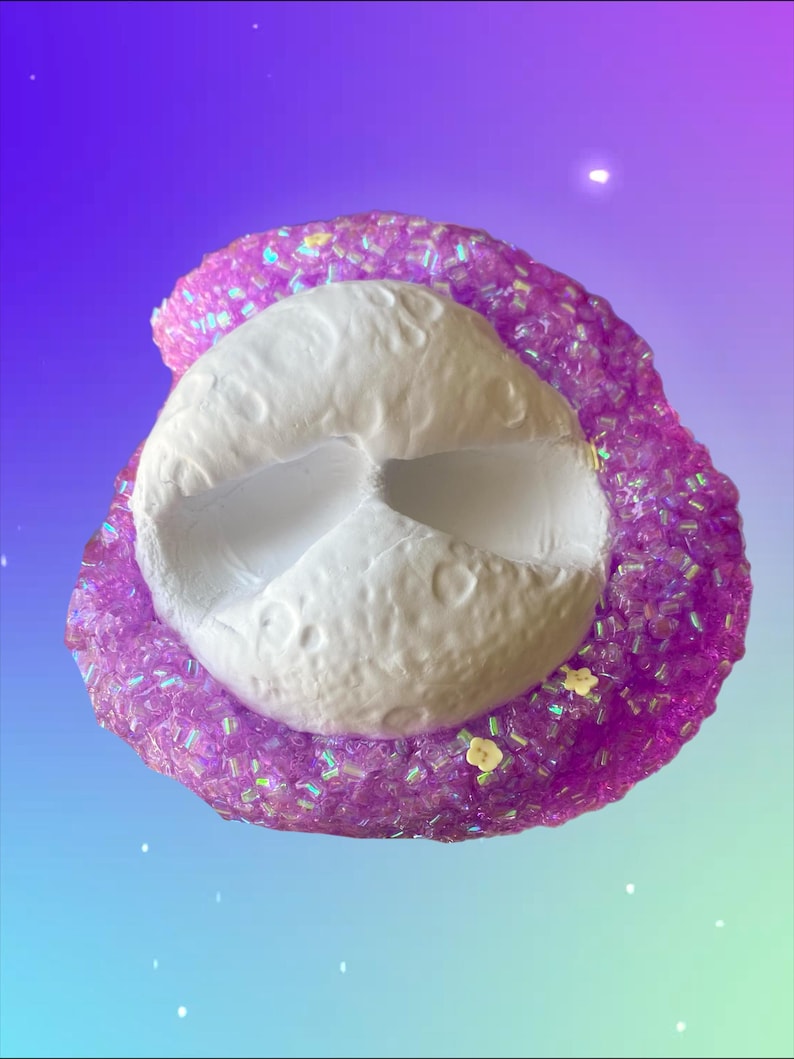 May include: A white, textured, moon-shaped object is surrounded by a sparkling, purple, glittery substance. The background transitions from purple to blue. Small yellow flower-shaped decorations are scattered around the object.