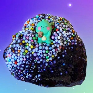 May include: A purple slime with a green cartoon character holding a cup of boba tea. The slime is covered in colorful sprinkles and stars.