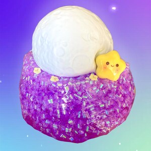 May include: A whimsical, handcrafted slime creation featuring a large, white, textured moon on top of a sparkling purple base. A yellow star with a smiling face is attached to the side, with small yellow flower accents.