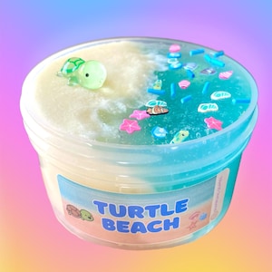 Pode incluir: Um recipiente de plástico transparente com slime com tema de praia. O slime é dividido em duas seções: uma amarelo claro e outra azul-esverdeada. A seção azul-esverdeada é decorada com pequenos encantos coloridos com tema marinho. O recipiente tem um rótulo que diz "Turtle Beach".