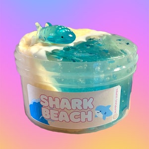 Könnte beinhalten: Ein durchsichtiger Plastikbehälter gefüllt mit blauem und weißem Schleim mit einer kleinen blauen Hai-Figur oben drauf. Der Behälter hat ein Etikett mit der Aufschrift "SHARK BEACH" und einer kleinen Hai-Grafik.