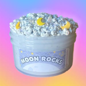 Puede incluir: Un recipiente de plástico transparente lleno de slime azul claro, cubierto con espuma blanca y decoraciones de media luna amarillas. El recipiente tiene una etiqueta que dice "Moon Rocks". El fondo es un degradado de rosa, morado y azul.