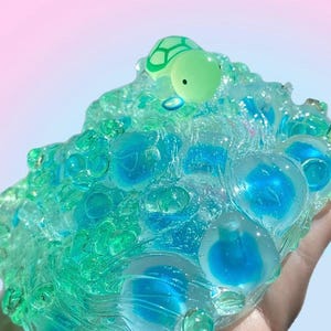 May include: A translucent, blue-green slime toy with embedded blue spheres and a small, green turtle figurine on top. The toy has a bubbly texture and is held in a hand, with a soft pink and blue gradient background.